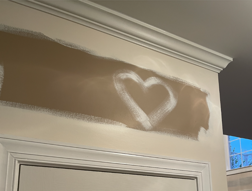 Image of a dark brown wall area above a doorway that has the first coat of a light beige paint along the edges. In the middle, there is a an outline of a heart, with the light beige paint used. 